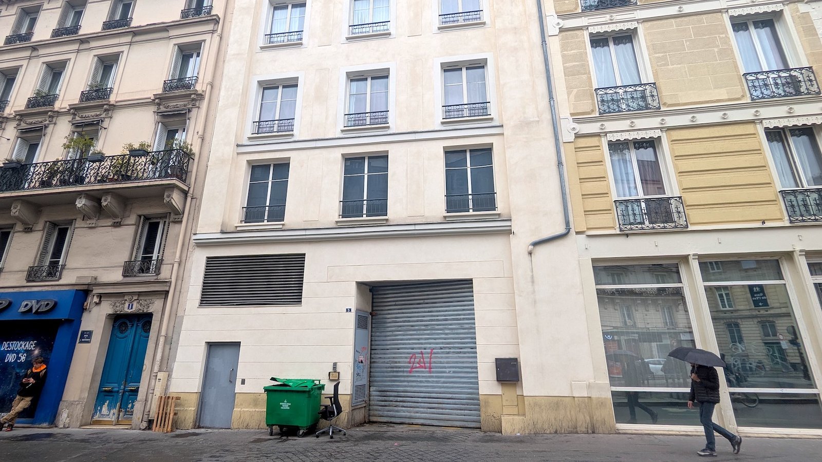 A man holding an umbrella passes in front of the partially fake house on 3 Rue de l'Aqueduc that has opaque windows on the first floor and normal ones on the other floors.